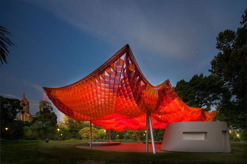 Dusk lighting transforms the canopy to glowing red above the cylindrical pavilion base in parkland