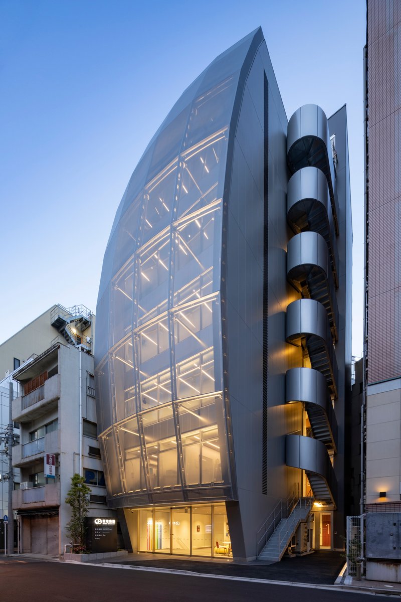 Street view of the faceted glass facade with illuminated structural frame and cylindrical stair tower at dusk