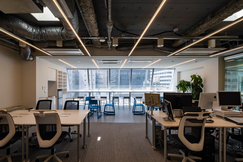 Open office floor with exposed ductwork, linear ceiling lights, and floor-to-ceiling windows overlooking neighboring buildings