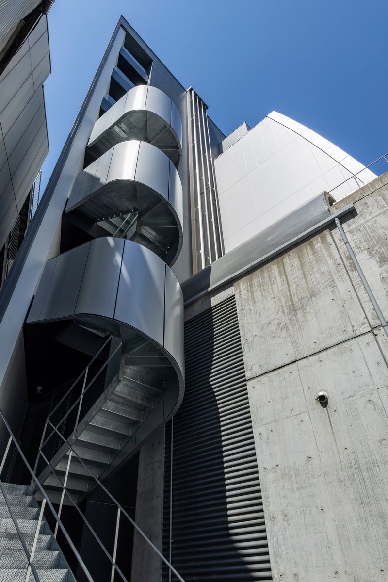 Exterior spiral staircase wrapping around a cylindrical glass tower beside a concrete wall