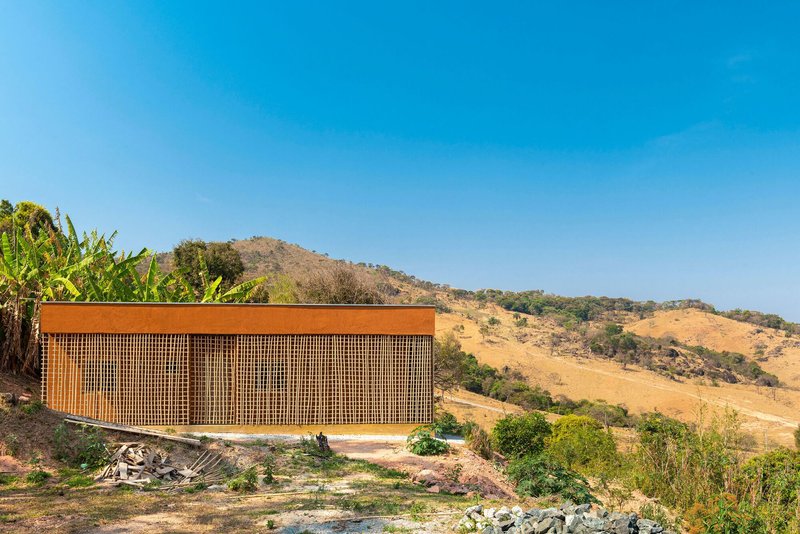 Elevated rectangular volume with timber lattice screen facade set against a dry hillside with banana plants