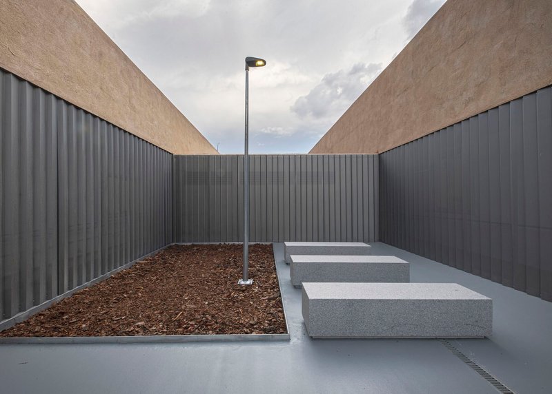 Enclosed courtyard with planted bed, concrete benches, and street lamp framed by ribbed metal walls