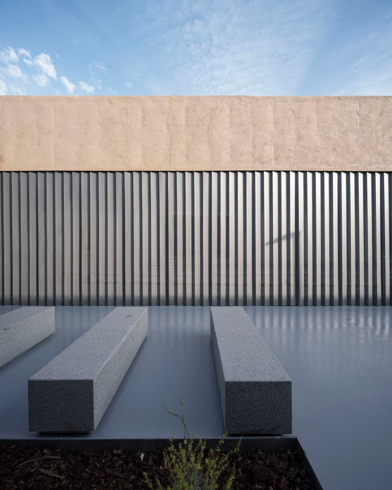Courtyard with grey concrete benches and vertical metal siding below a beige plaster parapet under blue sky