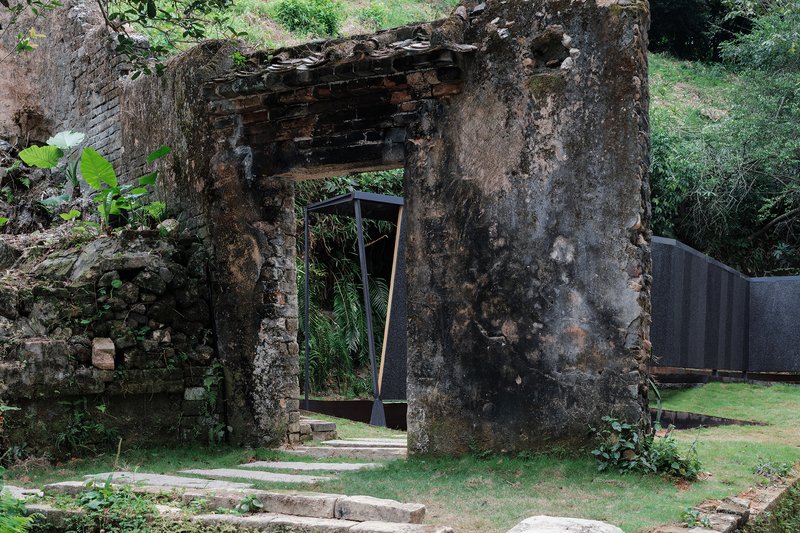 Crumbling stone ruin with a metal-framed door opening looking onto planted lawn and tropical growth