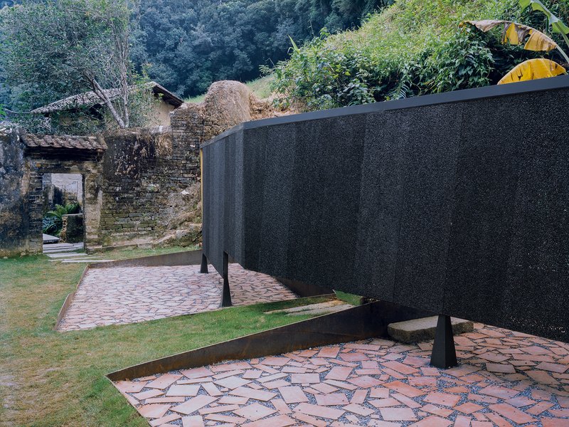 Curving black wall bisecting paved terraces with weathered stone ruins visible on the hillside behind