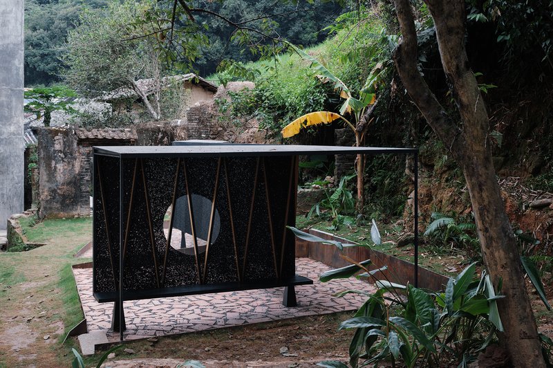 Pavilion with perforated black metal panels and circular opening set among banana trees and stone retaining walls
