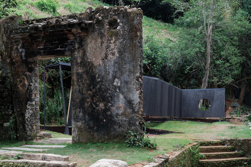 Weathered concrete ruins adjacent to dark curved wall with circular aperture in grass clearing