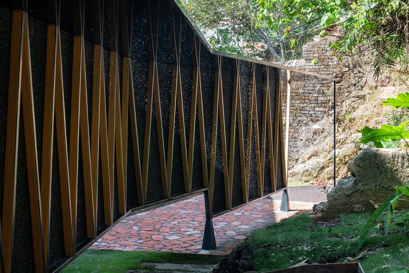 Long facade of terrazzo panels with timber slats and red tile floor leading toward stone ruins