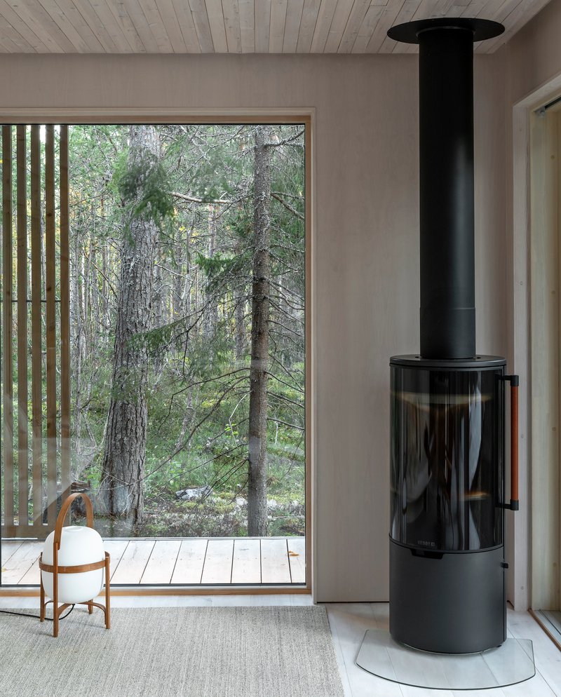 Freestanding black cylindrical wood stove beside a full-height window overlooking the forest