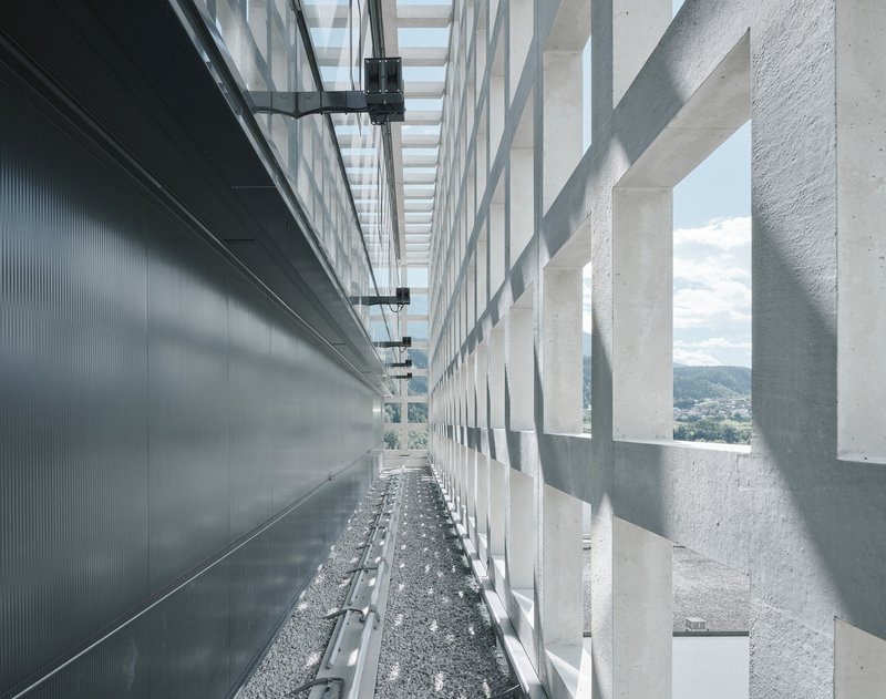 Narrow rooftop passage between metal wall panels and concrete light screen with gravel floor