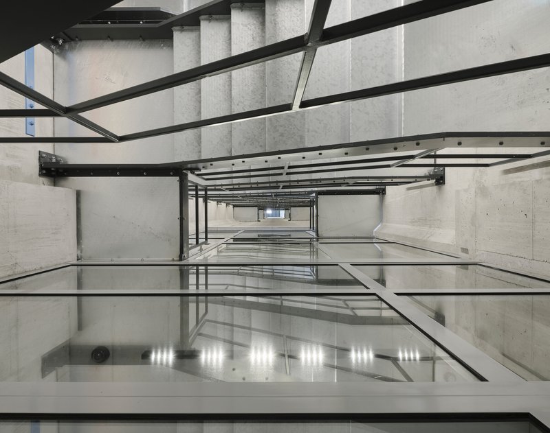 Looking down through a concrete elevator shaft with steel beams and reflective glass floor panels