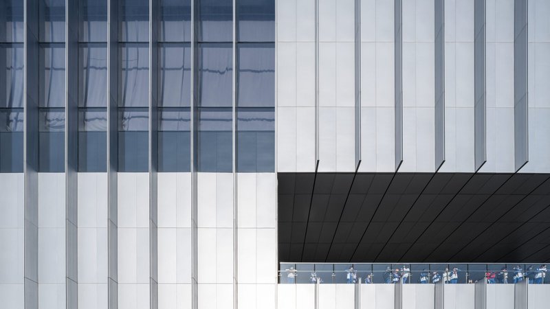 Facade detail showing white vertical panels meeting black soffit with glazed gallery strip revealing visitors inside