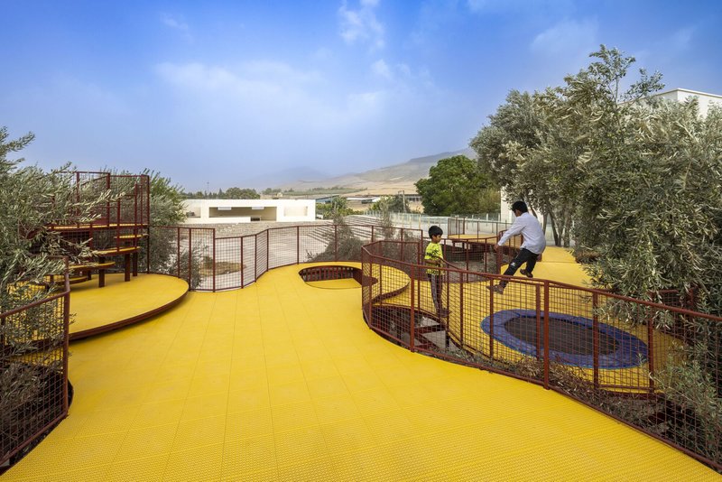 Yellow perforated deck with children playing along red mesh-enclosed walkways among olive trees