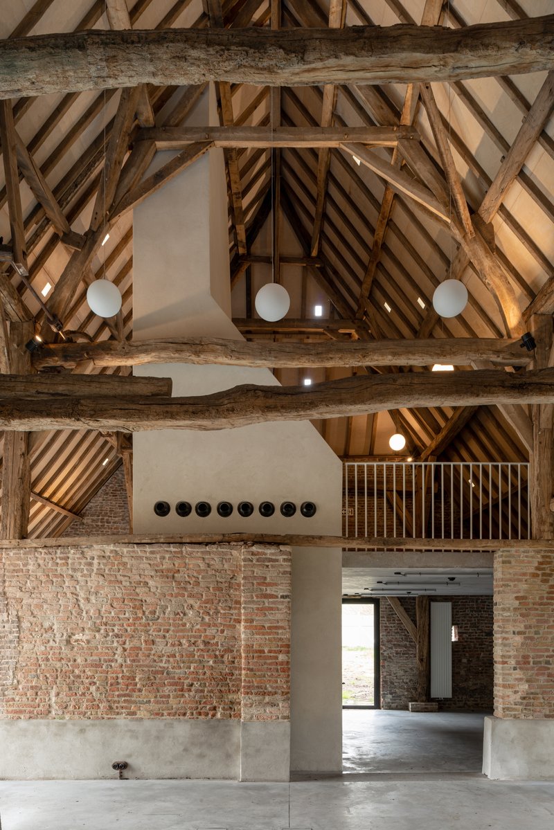 Double-height interior with exposed timber roof trusses, white plaster volume, and reclaimed brick walls under globe lighting