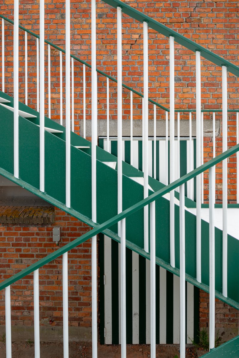 Green-and-white painted steel staircase with vertical bar railings ascending against exposed brick