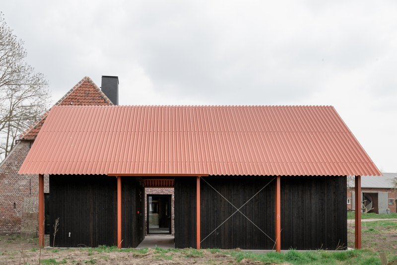 Black timber-clad barn volume with coral corrugated metal roof and orange steel columns under overcast sky