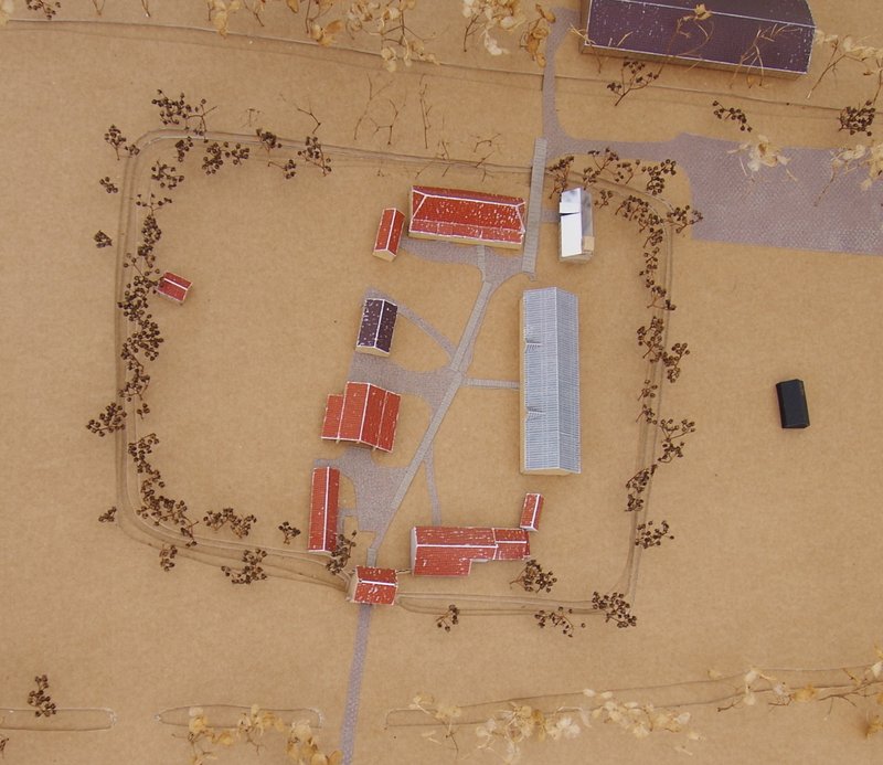Aerial view of farm buildings arranged along paths in a sandy clearing with scattered trees
