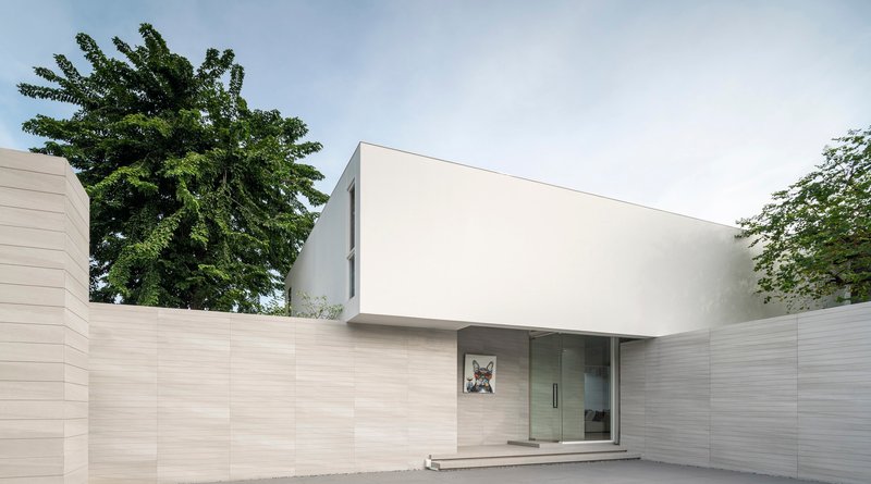 Street view of the white upper volume floating above horizontal stone walls and mature trees
