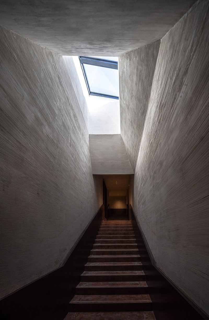 Timber staircase rising through a tall concrete light well with angled skylight above