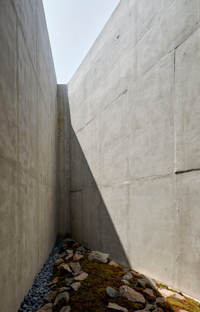 Narrow exterior courtyard between board-formed concrete walls with rocks and moss on the ground