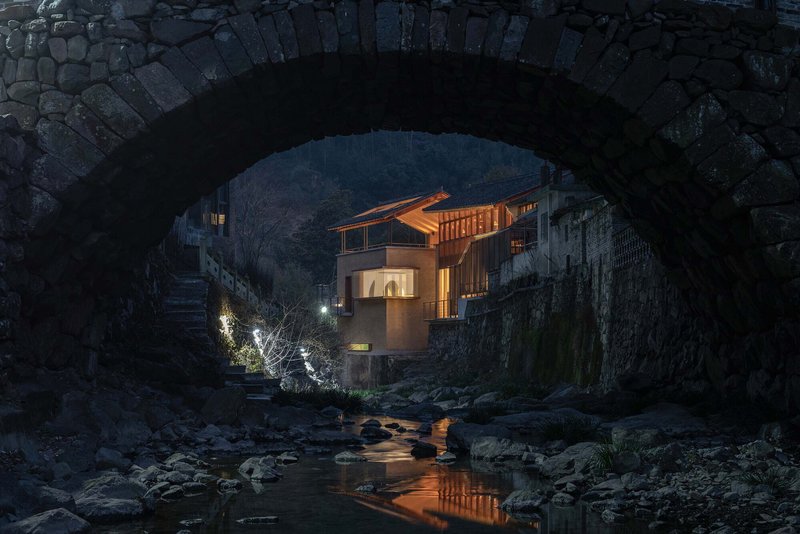 View through a stone arch bridge to the timber-clad facade illuminated at dusk above a rocky stream