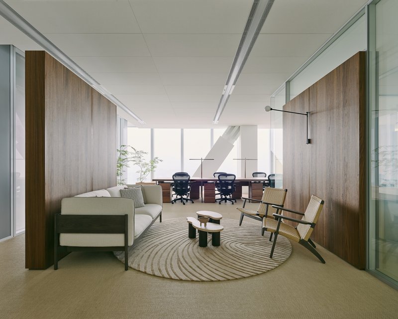 Office lounge area with walnut wall panels and circular rug separating seating from conference room beyond