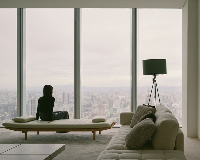 Person sitting on daybed by full-height windows overlooking the cityscape in soft afternoon light