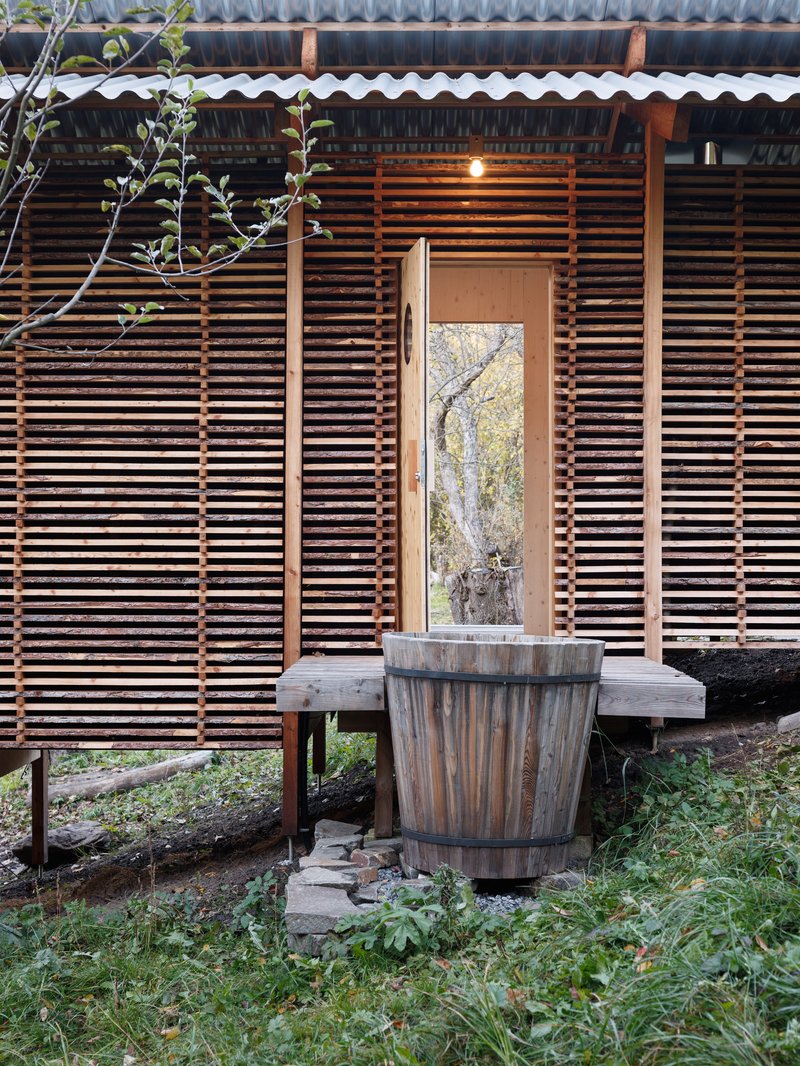 Facade with horizontal wood slats, tall narrow doorway, and wooden barrel on slatted deck at dusk