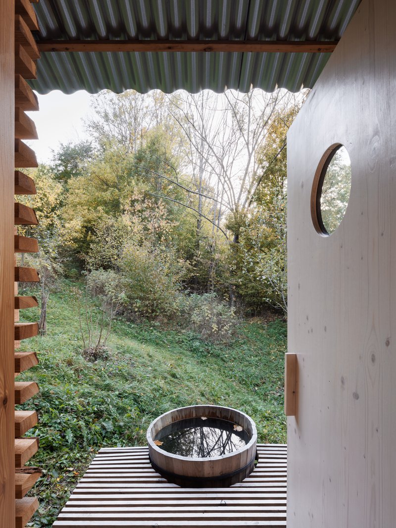 Covered deck with corrugated roof, plywood wall with circular window, and wooden tub overlooking autumn landscape
