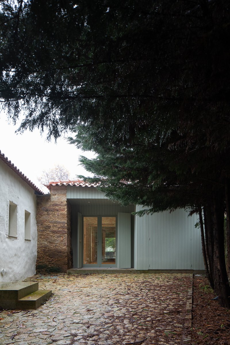 Corrugated metal pavilion inserted between whitewashed wall and stone structure under evergreen canopy