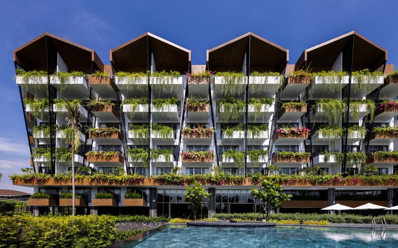 Facade with planted balconies and dark canopy roofs viewed across a swimming pool under blue sky