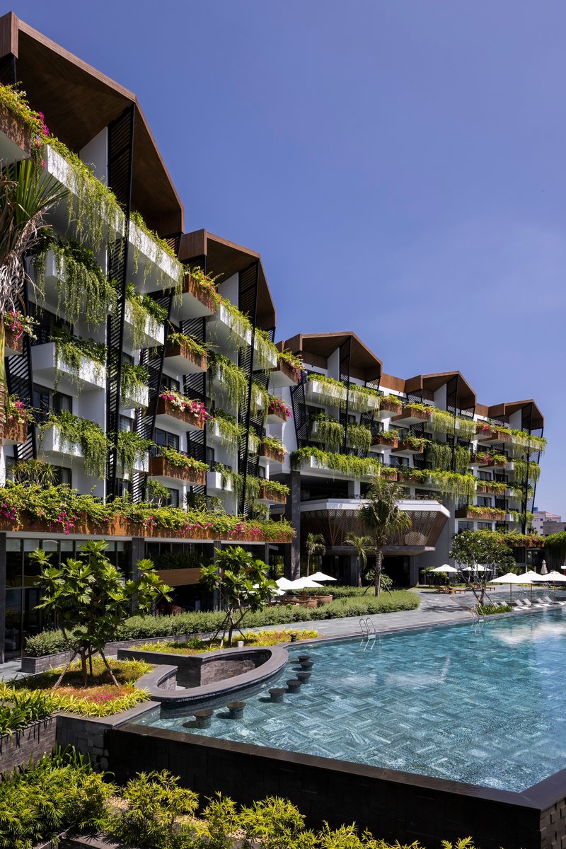 Planted facade rising above curved pool edge and walkway with young trees in afternoon light