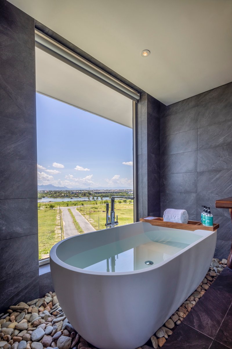 Freestanding bathtub on river stones against dark tile walls with a full-height window framing distant fields