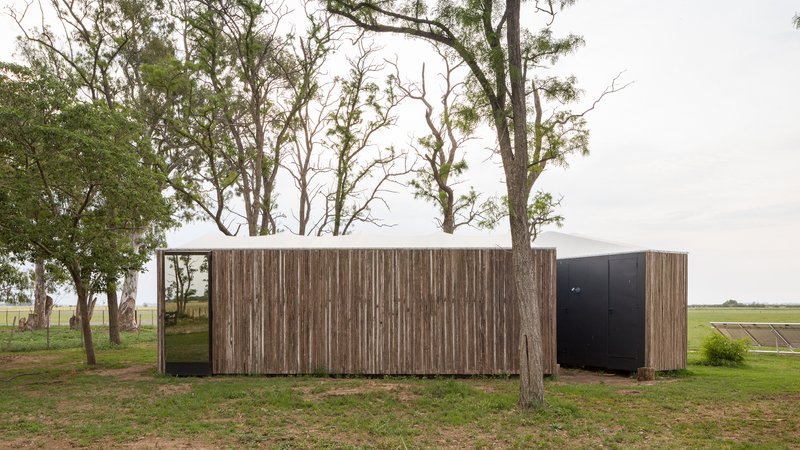 Timber-clad volume with black door nestled among mature trees in an open field