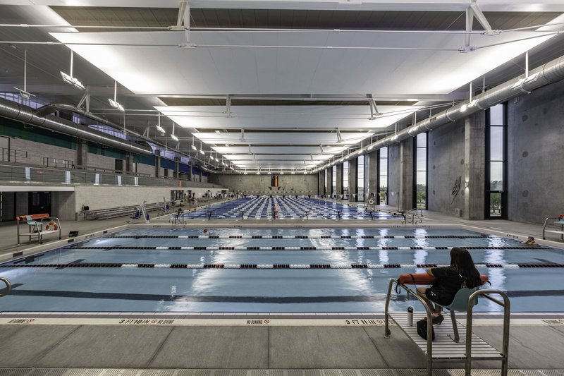 Olympic-length indoor swimming pool with concrete walls, exposed ceiling trusses and lane dividers