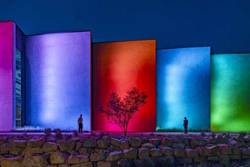 Colored illuminated concrete panels with two figures and a tree against a stone retaining wall