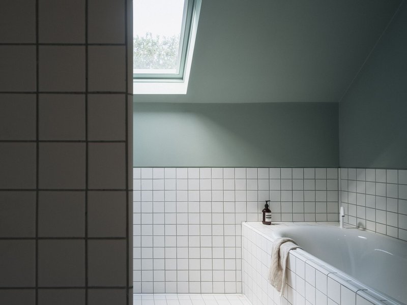Bathtub corner with white square tiles under a sloped ceiling and angled skylight casting soft daylight