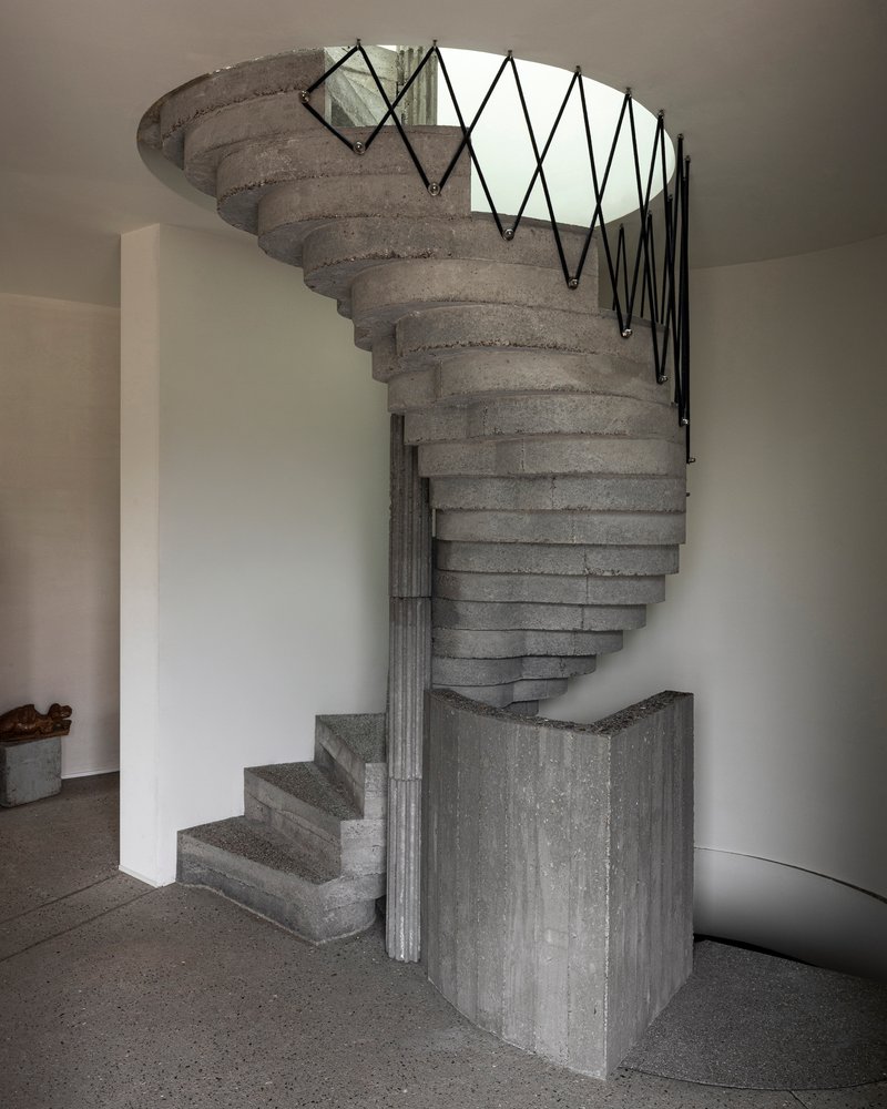 Helical concrete stair with zigzag metal railing and board-formed central column rising through circular opening