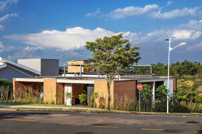Street view of the low-slung residence with brick walls, planted garden beds and rooftop terrace under scattered clouds
