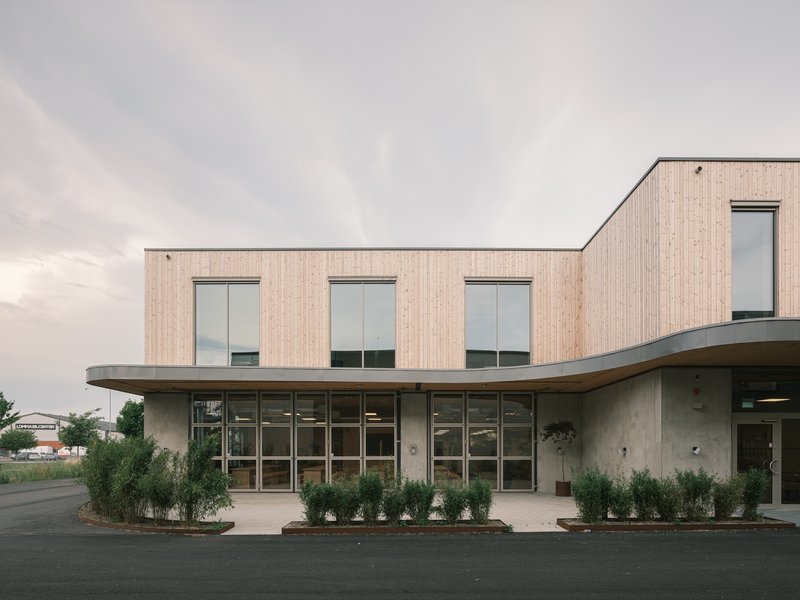 Two-storey facade with vertical timber cladding above a curved concrete base with planted beds at dusk