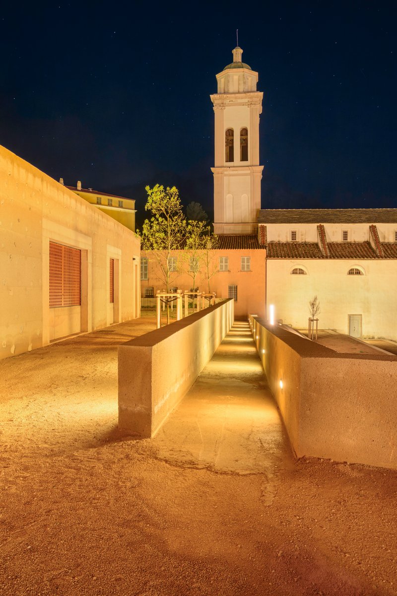 Night view of the illuminated courtyard passage with concrete walls framing the bell tower under a dark sky