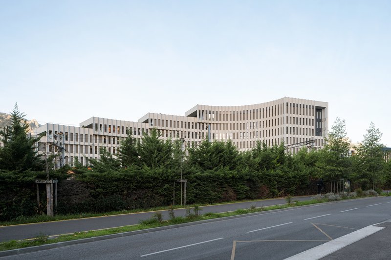 Street view of the curved building with ribbed facade behind roadside trees and hedges