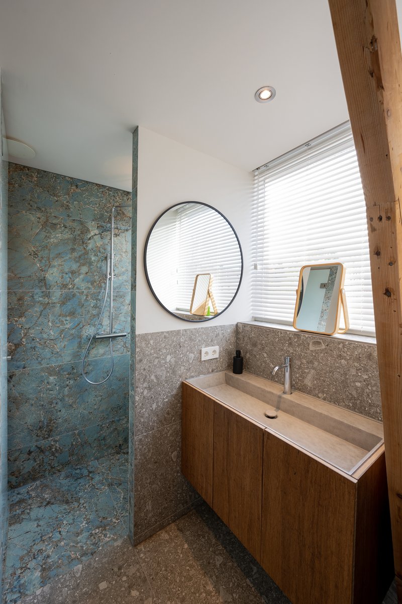 Vanity corner with round mirror, timber cabinetry and textured blue-green tile shower wall under natural light