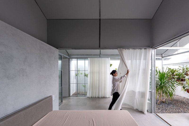 Double-height bedroom with concrete walls and person adjusting white curtain beside glazed courtyard