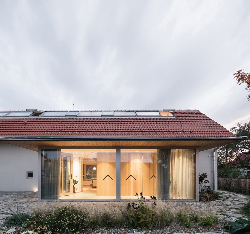 Full-height glazed facade with terracotta roof overhang illuminated at dusk beside flagstone terrace