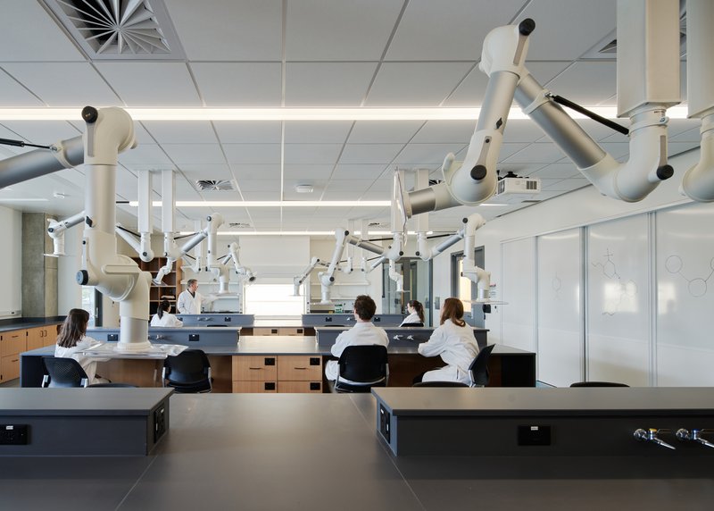 Laboratory workspace with rows of articulated microscope arms and students in white coats at workbenches