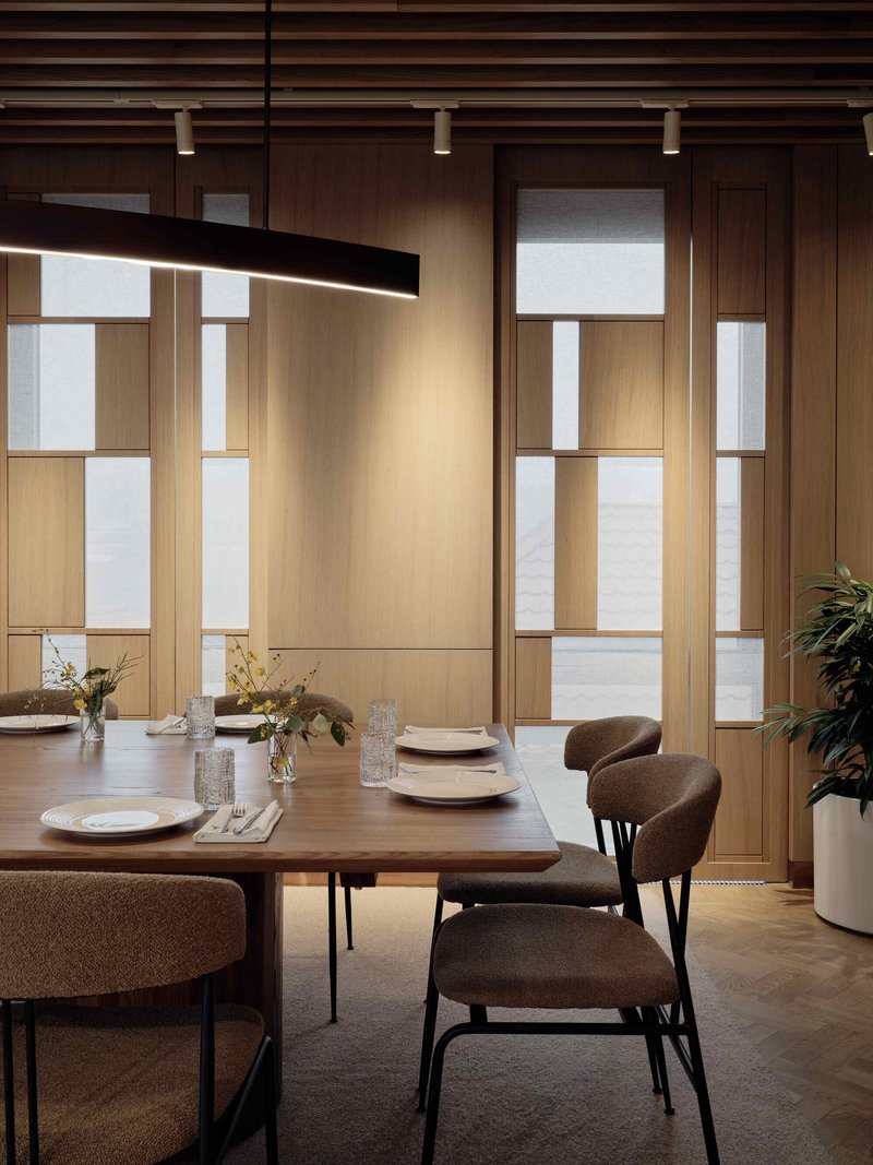Dining table beneath a linear black pendant with timber framed glass partitions in soft daylight