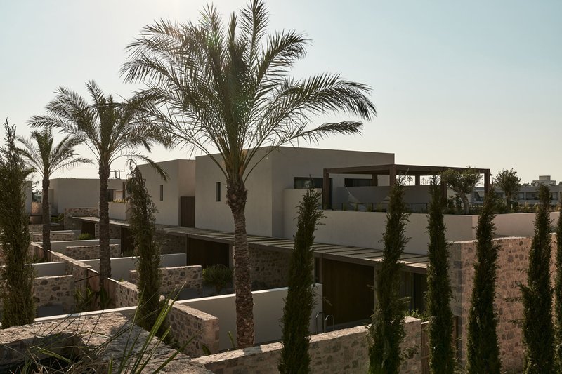 Clustered residential volumes with stone base walls and smooth stucco upper floors among cypresses and palms