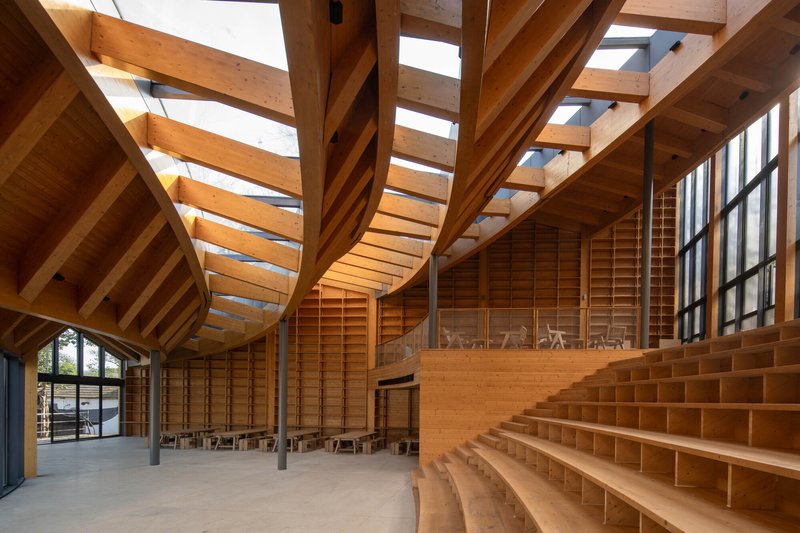 Interior assembly hall with curved timber beams, skylights, and tiered wooden seating along one wall