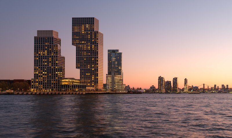Waterfront view of three staggered residential towers with illuminated windows at sunset